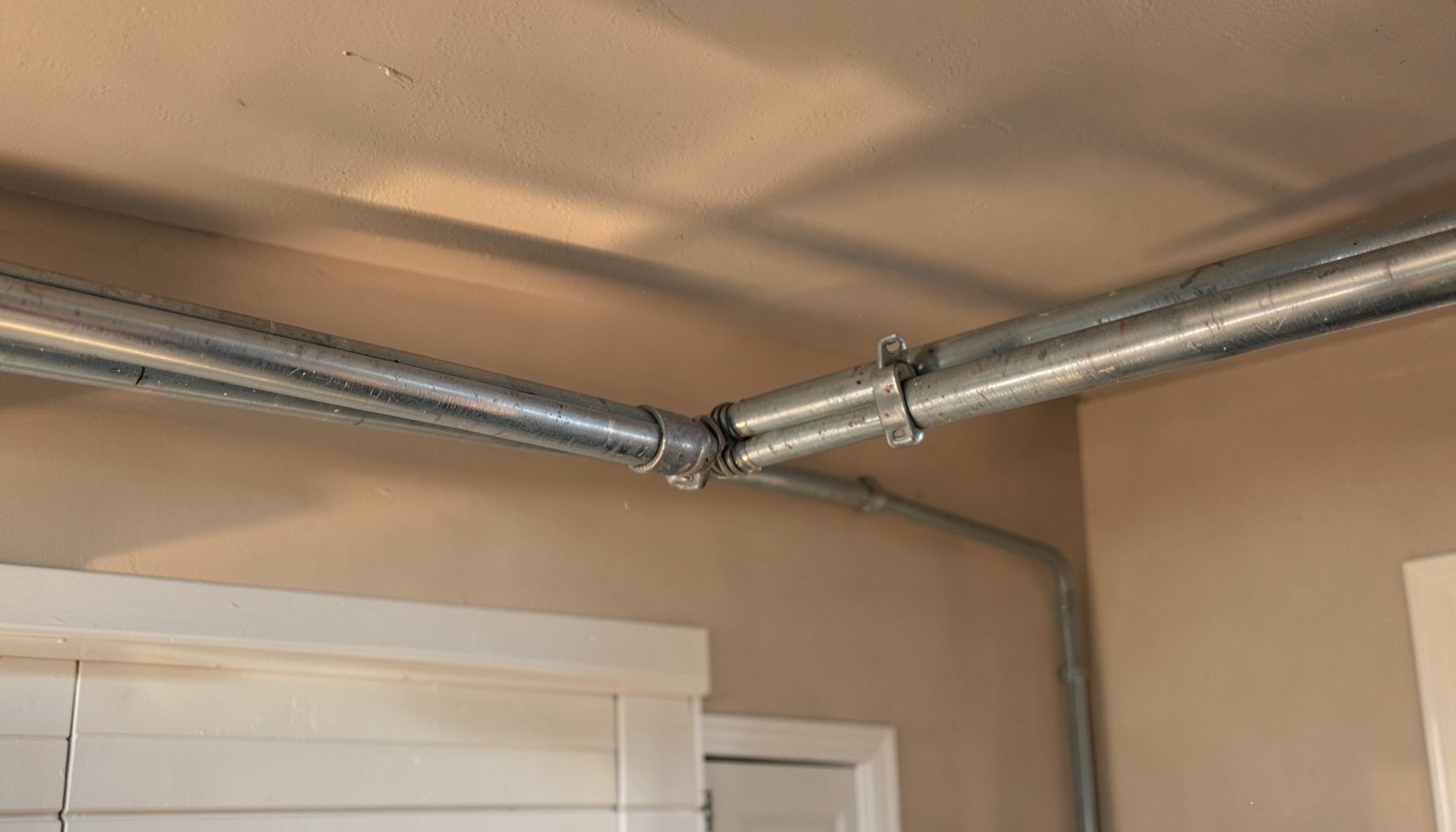 A close-up of two sturdy metal garage door springs mounted horizontally above a closed garage door, with warm afternoon light casting soft shadows across the track and ceiling. The scene feels calm and domestic, with neutral beige walls and a clean, well-kept garage interior.